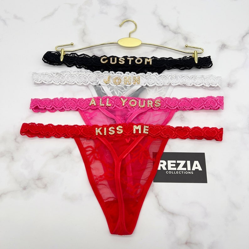 Może przedstawiać: Cztery stringi z czarnej, białej, r&oacute;żowej i czerwonej koronki, każdy z napisem ze złotych cyrkonii na pasku: CUSTOM, JOHN, ALL YOURS i KISS ME. Produkty są zawieszone na złotym metalowym wieszaku z metką REZIA COLLECTIONS.