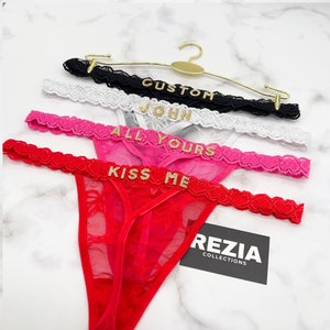 Może przedstawiać: Cztery stringi z czarnej, białej, r&oacute;żowej i czerwonej koronki, każdy z literami z cyrkonii tworzącymi napisy CUSTOM, JOHN, ALL YOURS i KISS ME. Złoty metalowy wieszak podtrzymuje przedmioty, a czarna karta zawiera napis REZIA COLLECTIONS.