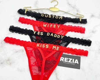May include: A display of four thong underwear pieces in black and red, each with a different gold-colored text: "CUSTOM", "WIFEY", "YES DADDY", and "KISS ME". The underwear is displayed on a gold hanger.