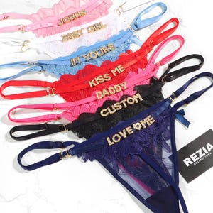 May include: A collection of lace thongs in pink, white, light blue, red, hot pink, black, and navy blue. Each features adjustable straps and gold-tone rhinestone lettering reading JOHNS, BABY GIRL, IM YOURS, KISS ME, DADDY, CUSTOM, and LOVE ME.