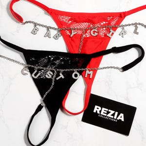 May include: Two thong underwear in red and black, adorned with silver chain accents and letter charms spelling "BABY GIRL" and "CUSTOM". The black thong features a lace design. A black business card with white text is also present.