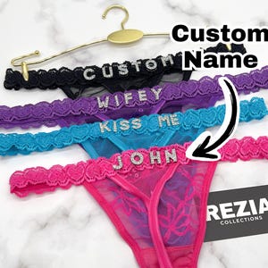 May include: Four lace thongs in black, purple, blue, and pink, each featuring a rhinestone-embellished waistband with the words CUSTOM, WIFEY, KISS ME, and JOHN. A gold-toned hanger holds the garments, and a black card displays REZIA COLLECTIONS.