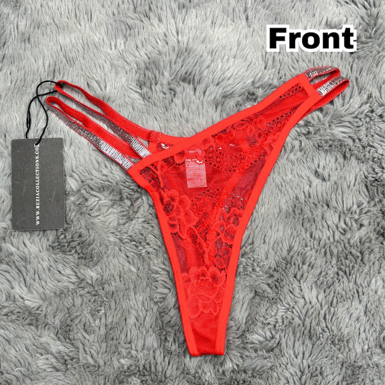 May include: A red lace thong featuring double side straps with silver glitter accents. The garment includes a black rectangular hang tag attached by a black string, with the text www.reziacollections.com printed in white on the tag.