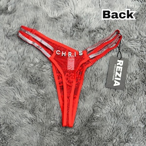 May include: A red lace thong featuring double side straps embellished with silver rhinestones and the name CHRIS spelled out in rhinestone letters across the front. A black tag attached to the side displays the text REZIA COLLECTIONS.