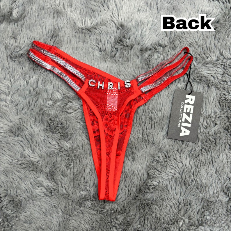 May include: A red lace thong featuring double side straps embellished with silver rhinestones and the name CHRIS spelled out in rhinestone letters across the front. A black tag attached to the side displays the text REZIA COLLECTIONS.