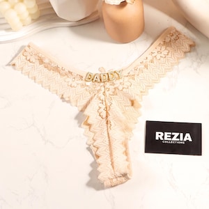 May include: A beige lace thong featuring a scalloped edge and a central gold-toned charm spelling the word DADDY in capital letters. A small black rectangular card with the white text REZIA COLLECTIONS is positioned to the right of the garment.