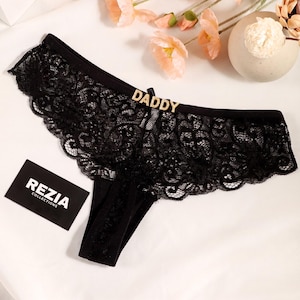 May include: Black lace underwear featuring a gold-toned metal charm that spells out the word DADDY centered on the waistband. The garment includes a small black tag with the white text REZIA COLLECTIONS printed on the front surface.