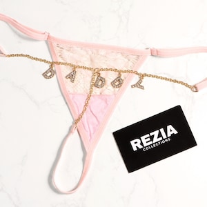 May include: A light pink thong featuring a lace front panel and thin elastic straps. A gold-toned chain is attached across the front, adorned with dangling rhinestone-encrusted letters spelling DADDY. A black card with the white text REZIA COLLECTIONS.