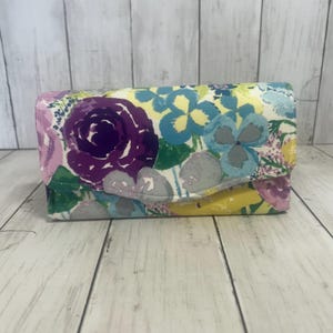 May include: A rectangular wallet featuring a vibrant floral pattern with large purple roses, light blue blossoms, and yellow accents on a white background. The wallet has a fold-over flap closure with a stitched edge, showcasing a colorful botanical print.