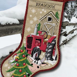 May include: A festive Christmas stocking with a cross-stitch design. It depicts a red tractor, a barn, and a cow, with the name "BRANDON" at the top. The stocking is trimmed with red rope and includes a Christmas tree and rooster.
