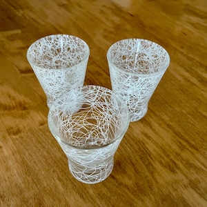May include: Three clear glass votive candle holders with a white web-like design. The candle holders are arranged on a wooden surface. The votives have a flared shape and are ideal for home decor.