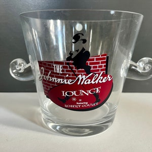 May include: A clear glass ice bucket with two handles. The bucket has a red, white, and black logo that says "The Johnnie Walker Lounge featuring Robert Goulet".