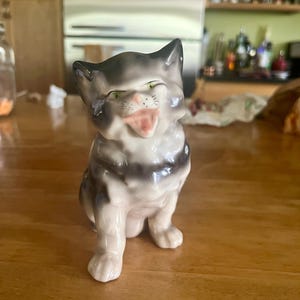 May include: A ceramic figurine of a cat in a sitting position, with a gray and white striped pattern. The cat has its mouth open, tongue out, and green eyes. The figurine is on a wooden surface.