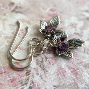 May include: Silver-toned floral earrings with purple gemstone accents. Each earring features a detailed leaf design and a faceted purple bead at the center. The earrings have a hook closure.