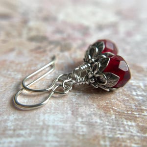May include: A pair of silver dangle earrings with faceted, deep red gemstone beads. The beads are encased in ornate silver floral settings. The earrings have a classic hook design.