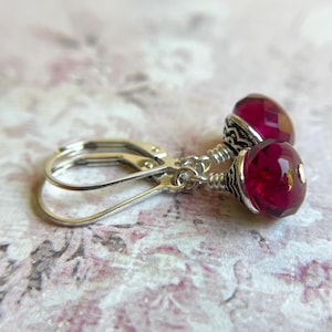 May include: A pair of leverback earrings featuring faceted, deep red glass beads. The beads are accented with silver-toned metal caps and wire wrapping. The earrings are displayed on a floral patterned surface.