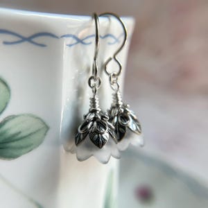 May include: A pair of drop earrings featuring frosted white bell-shaped flower petals encased in ornate, oxidized silver-toned metal caps with leaf detailing. The earrings hang from simple silver-toned metal hooks, creating a delicate floral design.