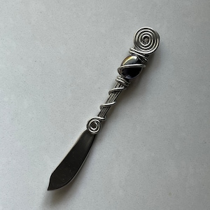 May include: A silver butter knife with a black bead and a wire wrapped handle. The handle is decorated with a spiral design.
