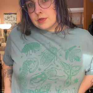 May include: A light blue t-shirt featuring a collection of illustrated insects in a teal color. The design includes various bugs, moths, and a snail, along with star-shaped accents. The shirt is a casual wear item.