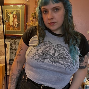 May include: A gray raglan t-shirt with black sleeves featuring a detailed black and white illustration of a bird with outstretched wings. The shirt is paired with black shorts and a black belt.