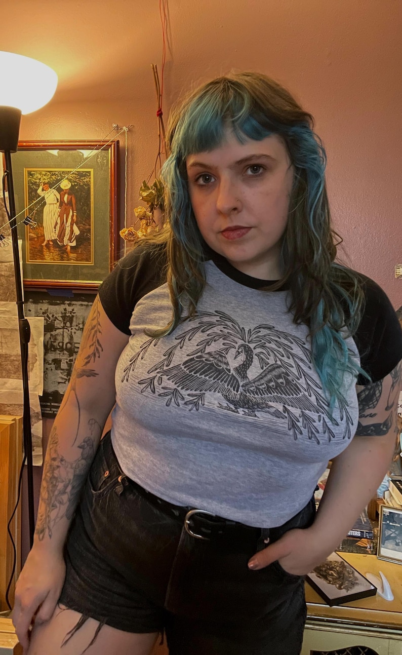 May include: A gray raglan t-shirt with black sleeves featuring a detailed black and white illustration of a bird with outstretched wings. The shirt is paired with black shorts and a black belt.