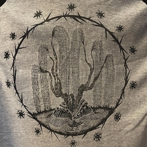 May include: A gray t-shirt featuring a detailed black illustration. The design includes a weeping willow tree with an eagle at its base, enclosed within a circle of thorns and stars. The artwork is centered on the shirt.