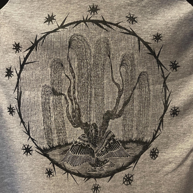 May include: A gray t-shirt featuring a detailed black illustration. The design includes a weeping willow tree with an eagle at its base, enclosed within a circle of thorns and stars. The artwork is centered on the shirt.