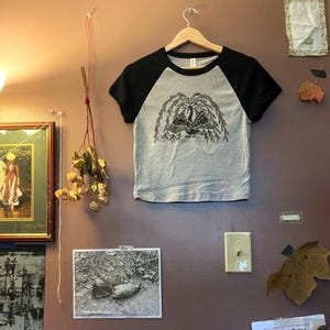 May include: A gray raglan t-shirt with black sleeves, hanging on a wooden hanger. The shirt features a black and white graphic of a weeping willow tree. The shirt is displayed on a brown wall with other decorative items.