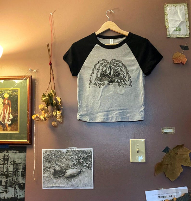 May include: A gray raglan t-shirt with black sleeves, hanging on a wooden hanger. The shirt features a black and white graphic of a weeping willow tree. The shirt is displayed on a brown wall with other decorative items.