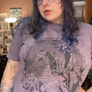 以下が含まれることがあります： 紫色のTシャツを着た人物。黒とグレーの馬とライダーのグラフィックが描かれています。丸いメガネをかけ、青い髪の毛が特徴です。シャツは黒いパンツと組み合わせています。