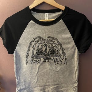 May include: A gray and black raglan crop top with a black and white illustration of two swans under a weeping willow tree. The shirt is hanging on a wooden hanger.