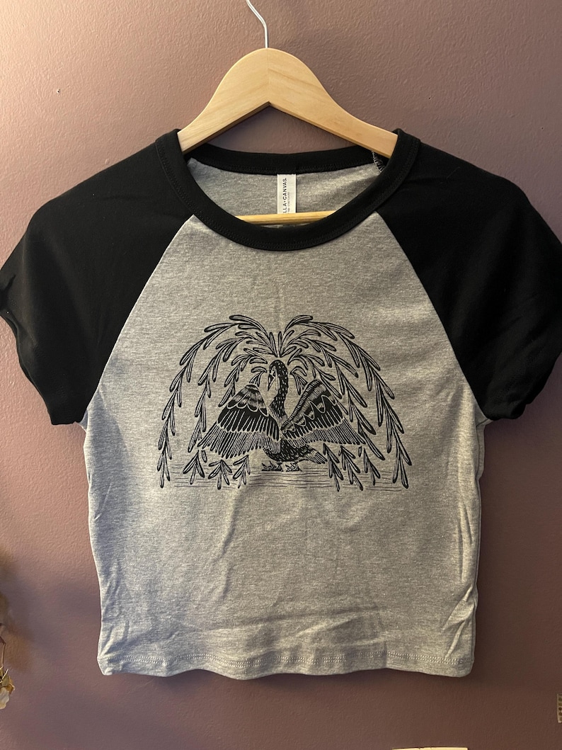 May include: A gray and black raglan crop top with a black and white illustration of two swans under a weeping willow tree. The shirt is hanging on a wooden hanger.