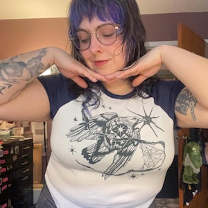 May include: White and navy blue raglan t-shirt with a black and white raven graphic holding a spider web. The raven has a star design on its chest. The shirt has short sleeves.