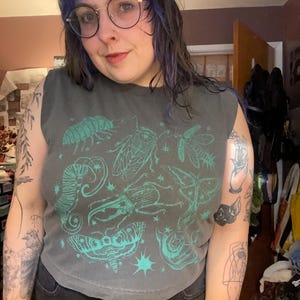 May include: A grey sleeveless top with a teal insect graphic. The design features various insects and starbursts. The top is worn with black denim jeans. The person in the image is wearing round glasses.