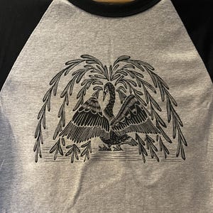 May include: A gray raglan t-shirt with black sleeves and a black collar. The shirt features a detailed black illustration of a swan with outstretched wings under a weeping willow tree design.