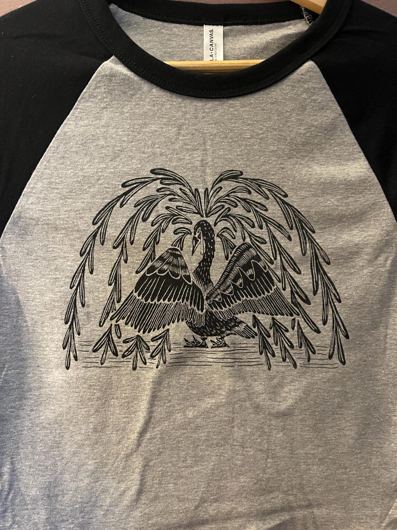 May include: A gray raglan t-shirt with black sleeves and a black collar. The shirt features a detailed black illustration of a swan with outstretched wings under a weeping willow tree design.