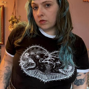 以下が含まれることがあります： 白の襟と袖トリムが付いた黒のリンガーTシャツ。シャツには、「LOUD BARK DEEP BITE」のテキストが入った、2匹の牙をむいたクマの白いグラフィックが描かれています。