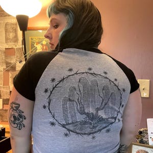 May include: Gray and black raglan baseball tee with a graphic illustration on the back. The illustration features a tree-like design within a circular frame of thorns and stars. The sleeves are black, and the body is gray.