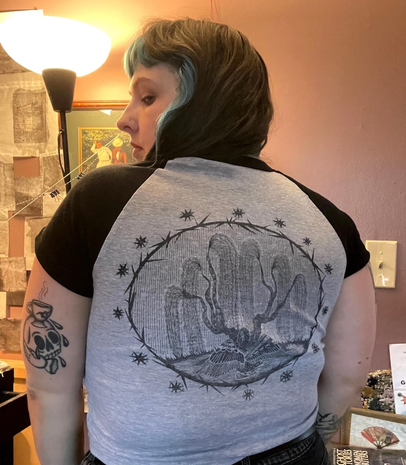 May include: Gray and black raglan baseball tee with a graphic illustration on the back. The illustration features a tree-like design within a circular frame of thorns and stars. The sleeves are black, and the body is gray.