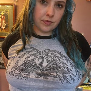 May include: A gray raglan t-shirt with a black collar and sleeves. The shirt features a large, detailed graphic of a bird with outstretched wings and foliage. The person in the image is wearing the shirt.