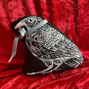May include: A black raven-shaped decorative pillow with a detailed white illustration of the bird's features. The pillow is tied with a light blue ribbon and set against a red velvet background. The design showcases a gothic or Halloween theme.