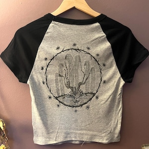 May include: A gray and black short-sleeved crop top with a graphic illustration on the back. The illustration is a detailed black and white drawing of a plant-like form within a circular frame, surrounded by star-like shapes.