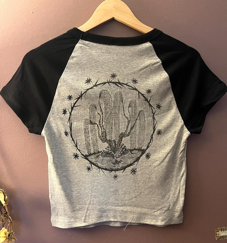 May include: A gray and black short-sleeved crop top with a graphic illustration on the back. The illustration is a detailed black and white drawing of a plant-like form within a circular frame, surrounded by star-like shapes.