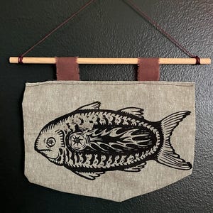 May include: A decorative wall hanging featuring a stylized black fish design on a gray fabric banner. The banner is suspended from a wooden dowel with maroon fabric accents and a maroon string for hanging. The fish has a compass-like design.