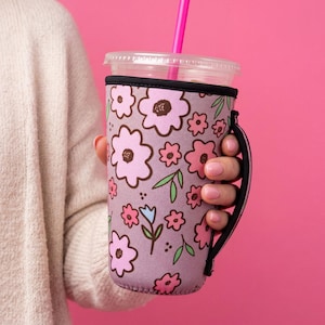 May include: A lavender neoprene beverage sleeve featuring a repeating pattern of pink and light pink flowers with dark brown centers, green leaves, and small blue accents. The sleeve includes a black fabric handle and a clear plastic lid with a pink straw.