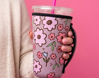 May include: A lavender neoprene beverage sleeve featuring a repeating pattern of pink and light pink flowers with dark brown centers, green leaves, and small blue accents. The sleeve includes a black fabric handle and a clear plastic lid with a pink straw.
