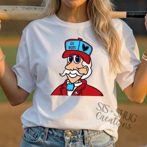 May include: White t-shirt featuring a cartoon character wearing a red hat with the words "GO REBS" and a black heart. The character has a white mustache, a red jacket, and a blue bow tie. The text "SIS + SHUG Creations" is also visible.