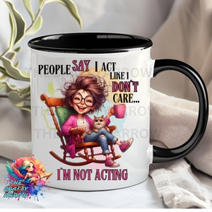 May include: White ceramic mug with a black rim and handle. The mug features a cartoon illustration of a woman sitting in a rocking chair with a cat. The text on the mug reads "People say I act like I don't care... I'm not acting."
