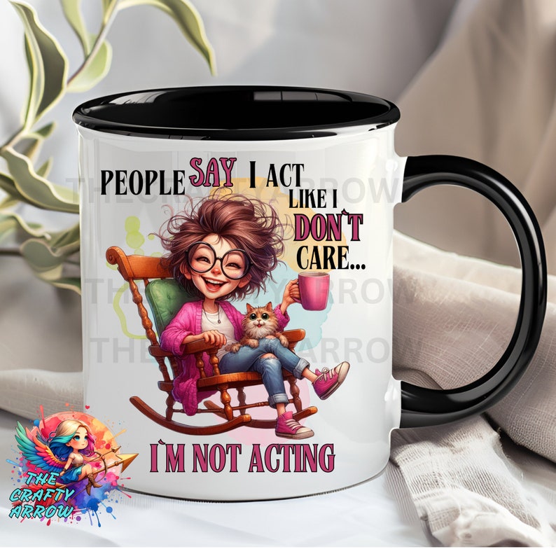 May include: White ceramic mug with a black rim and handle. The mug features a cartoon illustration of a woman sitting in a rocking chair with a cat. The text on the mug reads "People say I act like I don't care... I'm not acting."