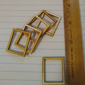 May include: Small, unfinished wooden square frames in various sizes, with a wooden ruler showing inches. The frames are light brown with a slightly darker border. The frames are scattered on a lined paper surface.